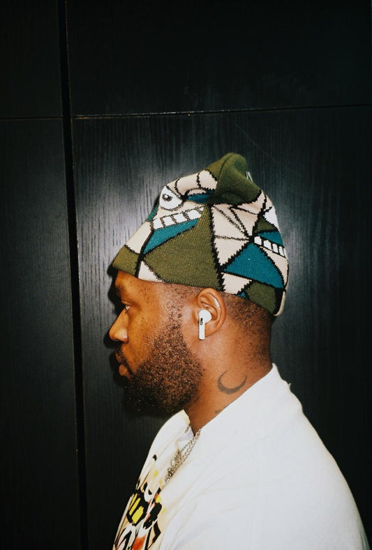 A man in profile wearing a knit beanie with a geometric pattern in olive green, teal, cream, and black, paired with a white t-shirt and white wireless earbuds, set against a dark wood-paneled background.