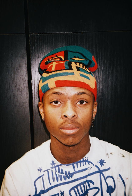A close-up portrait of a young man wearing a colorful knit beanie with a geometric pattern in teal, red, beige, and black, paired with a white graphic t-shirt against a dark paneled background.
