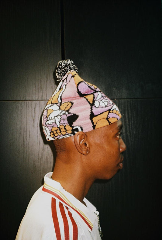 A side-profile portrait of a man wearing a pink knit beanie with a multicolor cartoon graphic print and a black-and-white pom-pom. He is wearing a white shirt with red stripes against a dark paneled background.