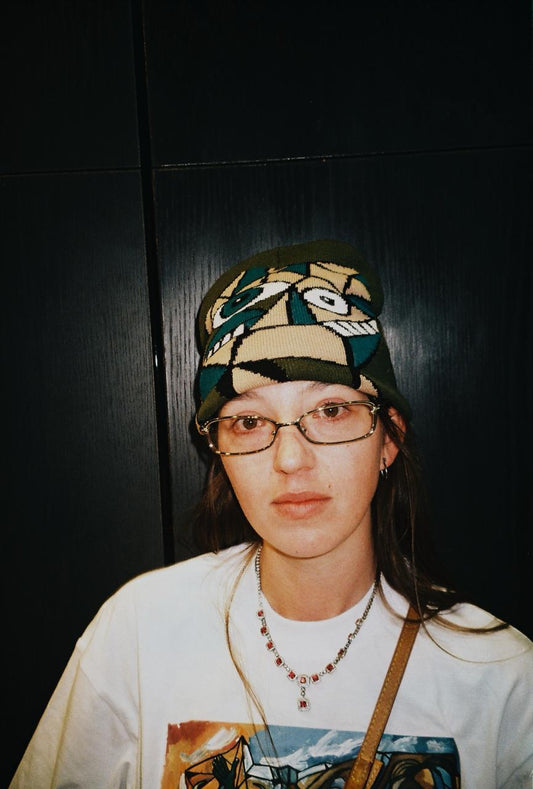 A person wearing a green knit beanie with a stylized face pattern, glasses, a white graphic t-shirt, and a necklace, standing against a dark paneled background.