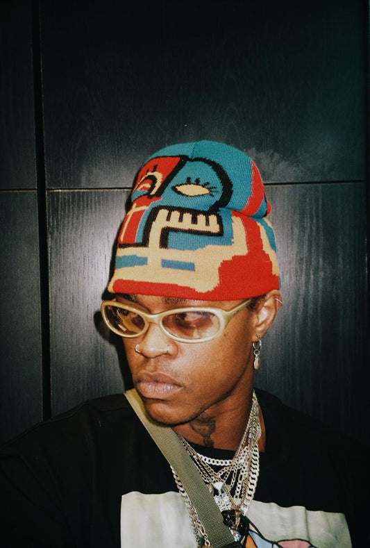 A close-up portrait of a person wearing a colorful knit beanie with a stylized face design, gold-rimmed glasses, layered silver necklaces, and a black shirt against a dark paneled background.